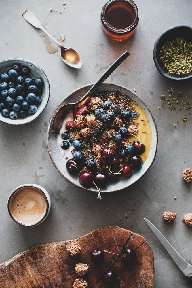 Oat granola yogurt bowl with cherries, blueberries, honey, nuts, coffee, wooden board in foreground
