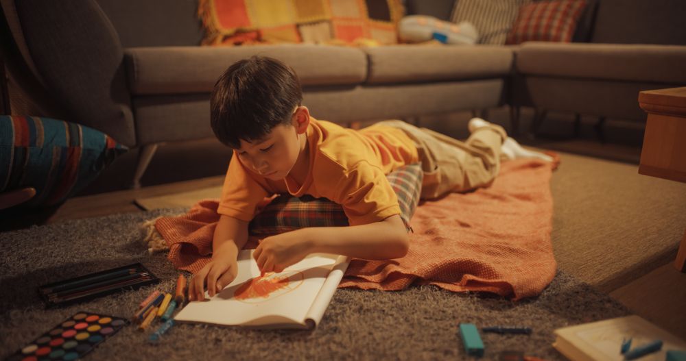 Cute Korean Boy Laying On The Floor And Drawing In Notebook With Pens, Pencils