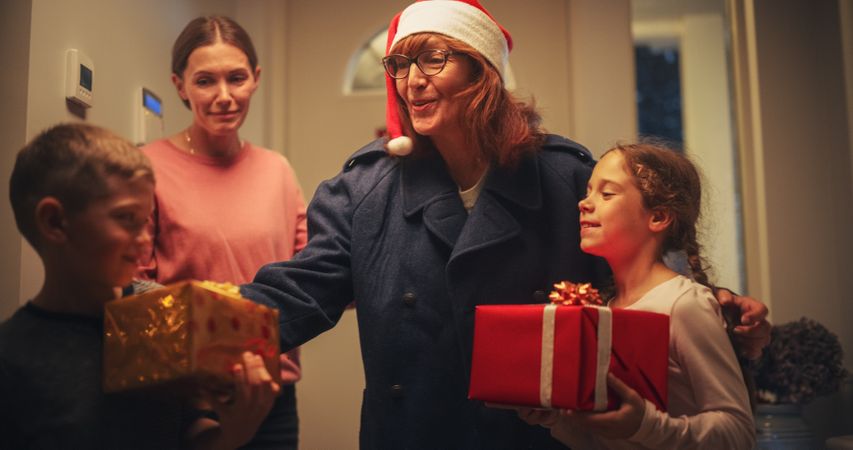 Young Mother Meeting Grandparent by the Door in a Decorated Home with Garlands, Small Boy and Girl Running to Meet Their Grandmother with Christmas Gifts and Presents