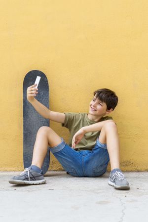 Boy sitting on ground leaning on a yellow wall and taking a selfie, vertical