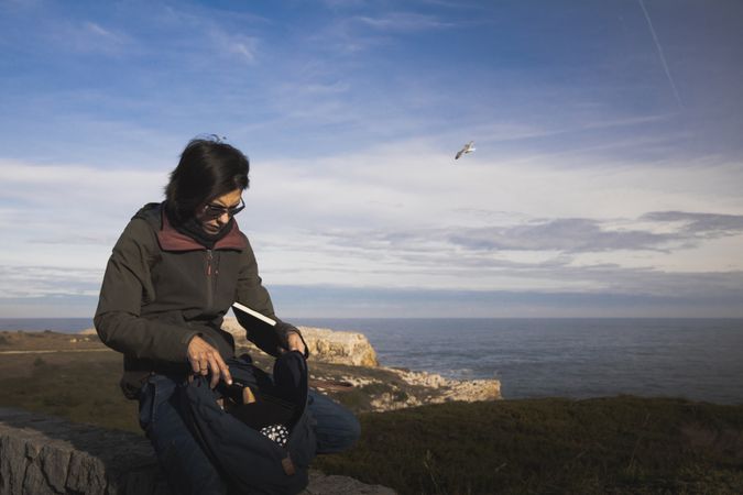 elegant woman taking out a book from back pack and sitting on meadow near beacon on shore and wonderful sky