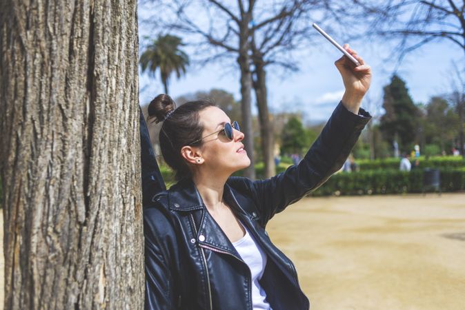 Side view of a young laughing hipster woman standing and leaning on a tree at park in sunny day while using a mobile phone to take a selfie