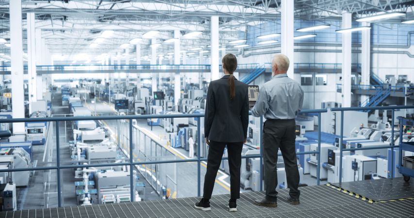 Male Engineer and Female Project Manager Standing on a Platform, Talking, Using Laptop Computer and Monitoring Production at a Modern Automated Electronics Manufacture with AI Robotic Arms