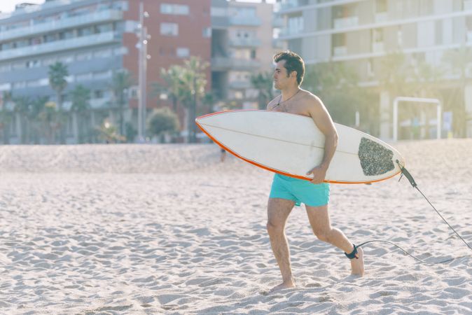 Male surfer walking on beach with board attached to his leg with leash on sunny day