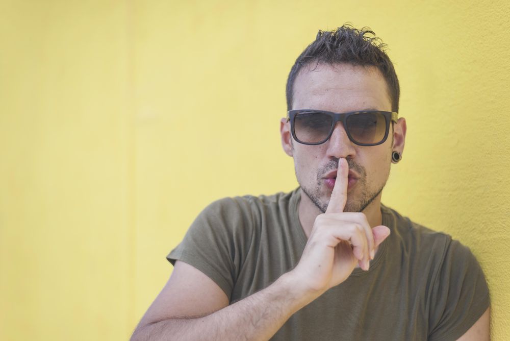Male making “quiet” sign in front of yellow wall outside