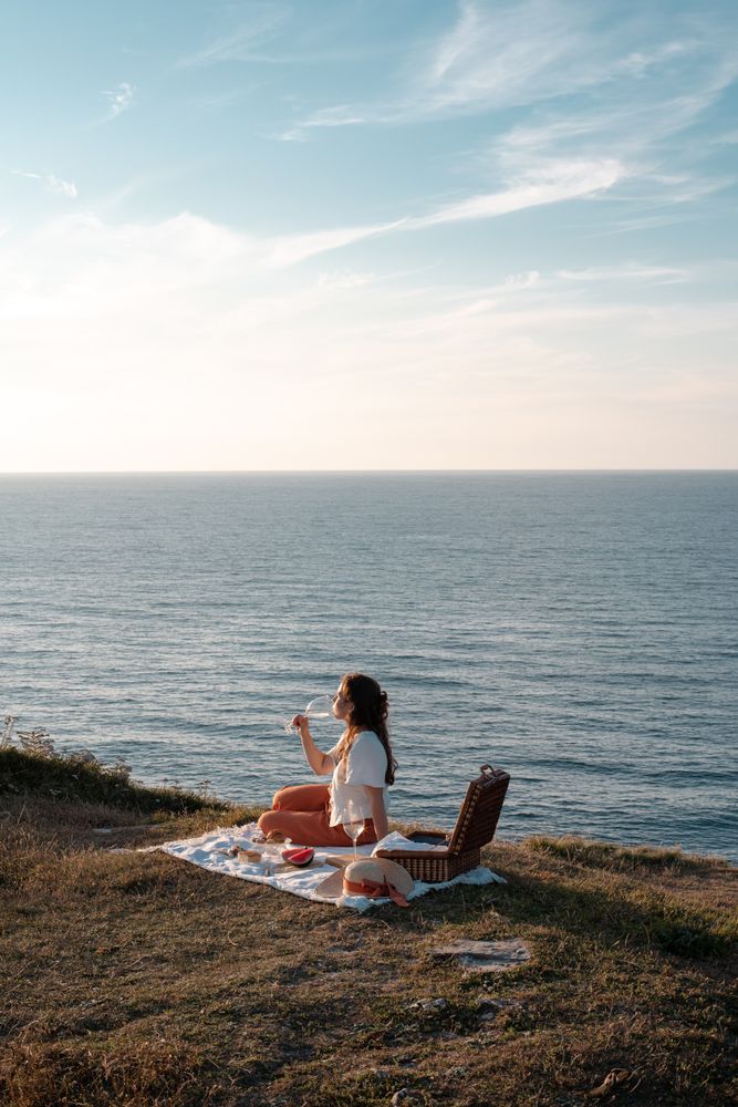 Woman with glass of drink on picnic mat looking at sea and mountains