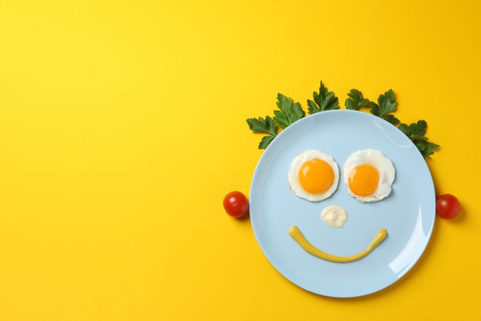 Blue plate with smiley face on it made of eggs and condiments, with vegetable hair, copy space