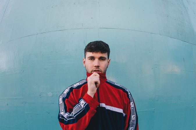 Young white male model zipping up track jacket over chin against blue wall