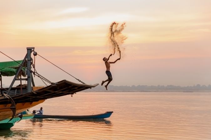 Boy jumping into water from a boat throwing sand in the air at sunset