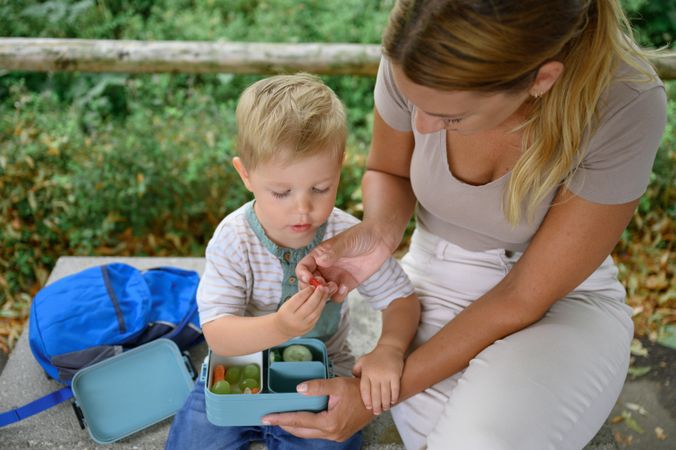 Crop young mother and son sitting on bench with lunchbox in daylight