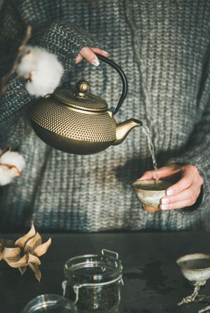 Woman in cozy sweater pouring from traditional Japanese tea set, with dried cotton