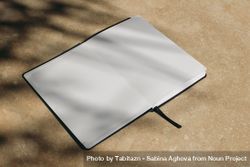 Tropical summer mockup. Open diary, book with black cover in sunlight. Blank pages. Harsh tree branches shadows. Beige marble stone texture, background. Holiday template. Flat lay, top view, no people 4d8XYA