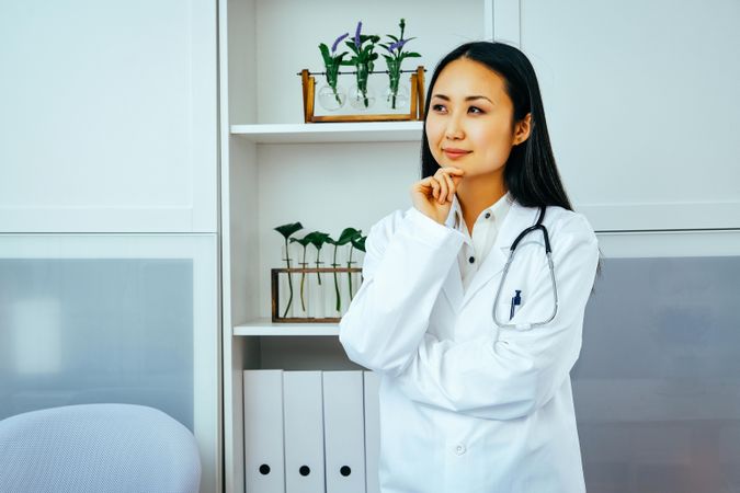 Contemplative Asian doctor thinking about something in her office with hand to her chin