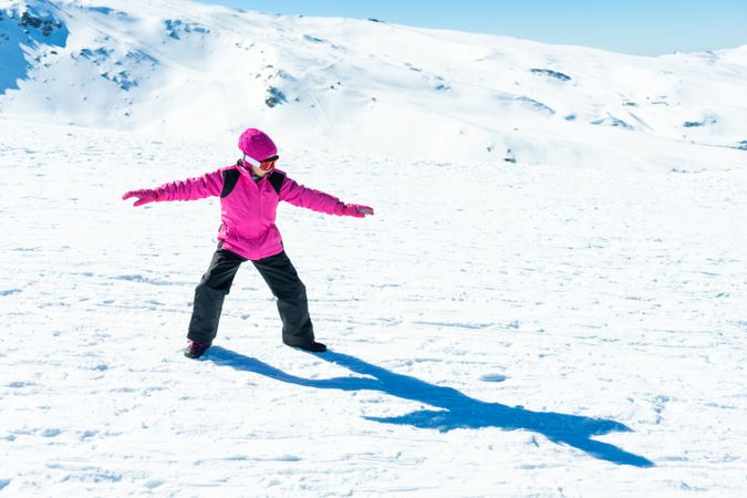 Child in pink snow suit practicing snow board poses on snowy hill