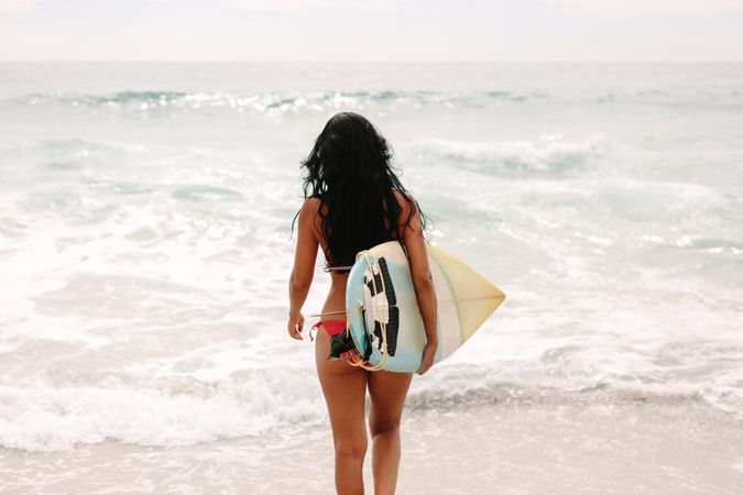 Rear view of woman in bikini with surfboard walking toward sea