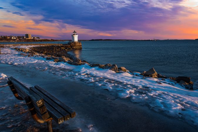 Wooden bench near lighthouse on rocky seashore at sunset