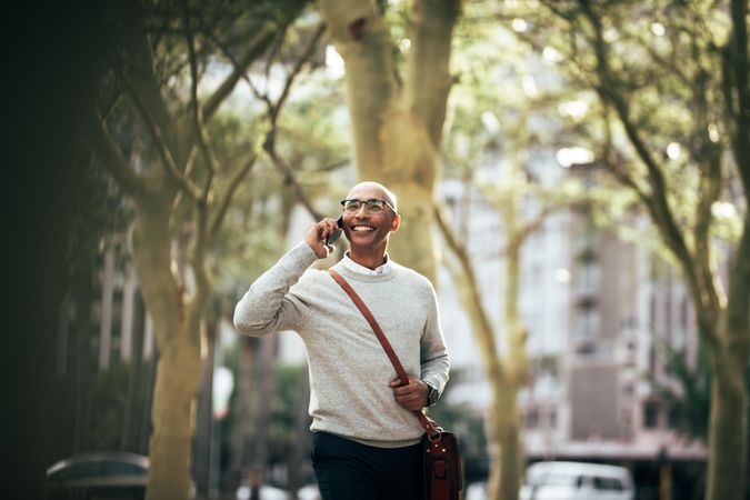 Smiling businessman talking over cell phone while walking on city street to work
