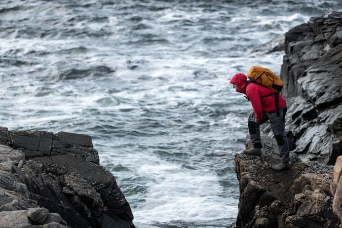 Person in red jacket and backpack on rocky cliff beside ocean