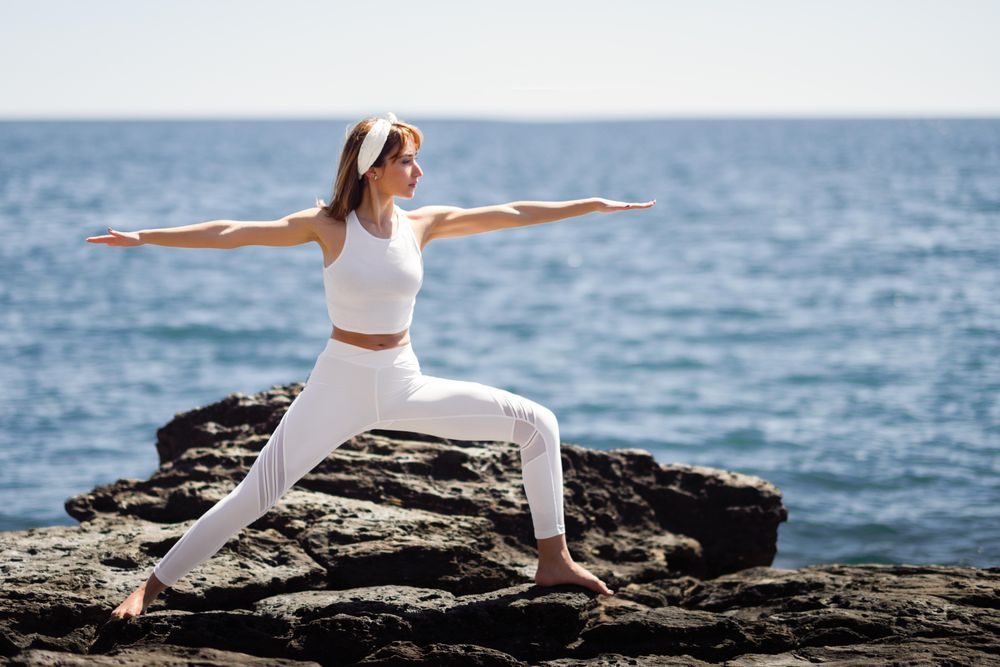 Woman wearing sport clothes and doing warrior B pose on rocks on the coast