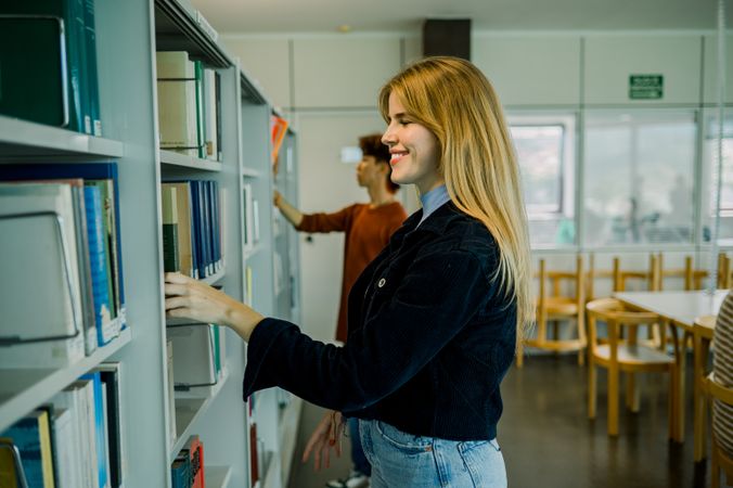 Cheerful woman standing near bookshelf and choosing book in library