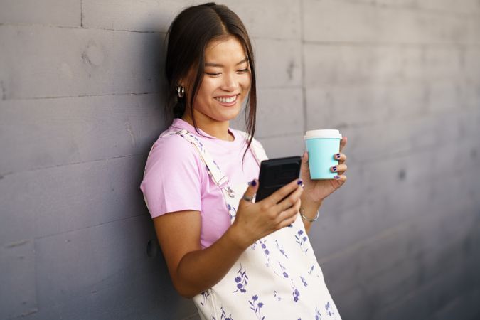 Happy Asian female wearing casual clothes holding phone while leaning against wall with coffee