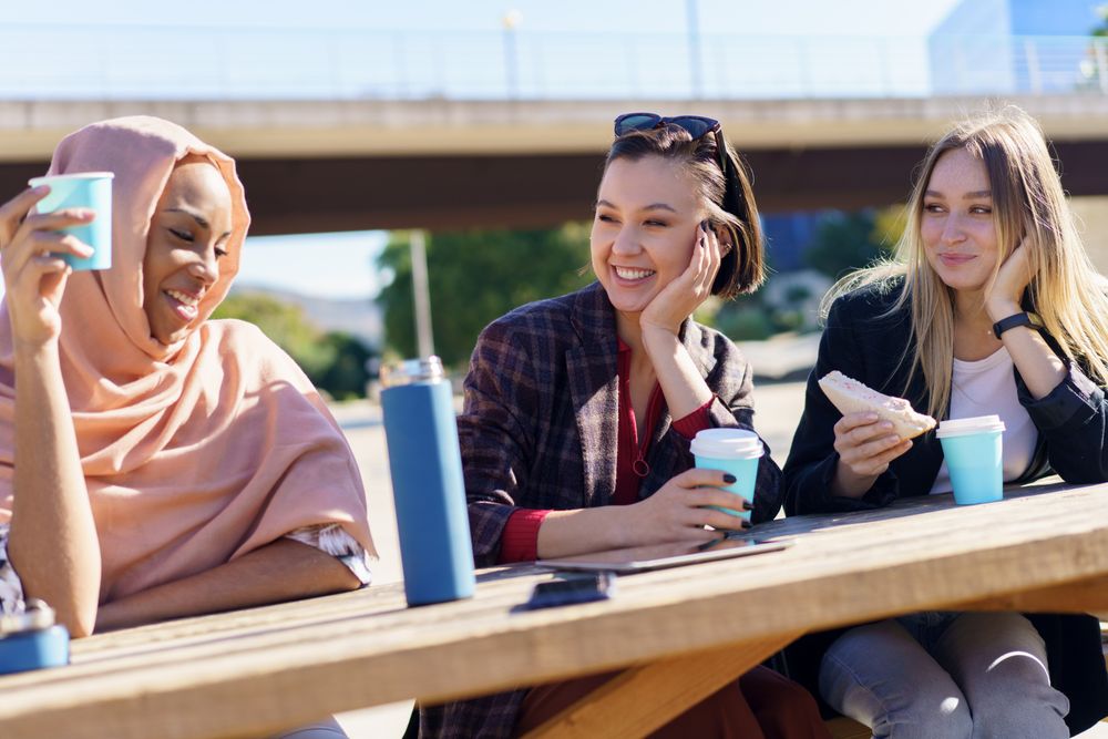 Smiling female friends sitting outside with warm drinks and sandwiches