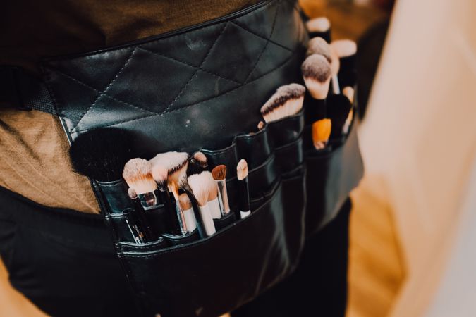 Unrecognizable makeup artist with brushes in apron bag