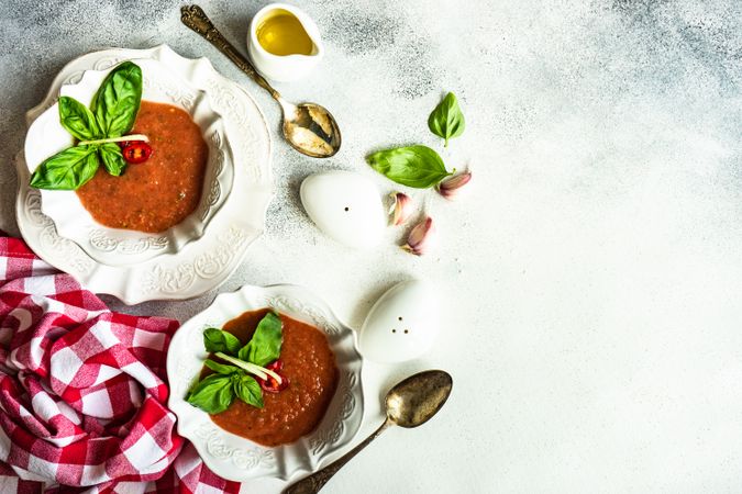 Two bowls of traditional gazpacho soup with basil, garlic, and salt and pepper shakers