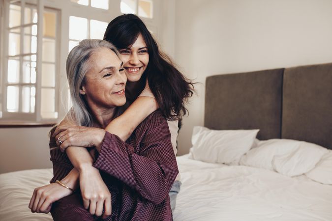 Older mother sitting on bed with her adult daughter hugging her from behind and holding her hands