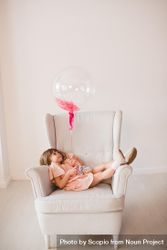Girl in pink dress holding a balloon lying on gray armchair bEJVG0