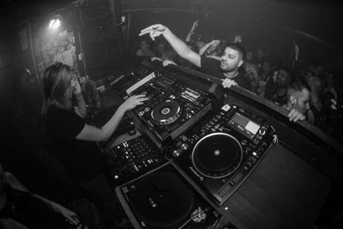 Fisheye shot of man reaching up into DJ booth as female DJ plays at nightclub