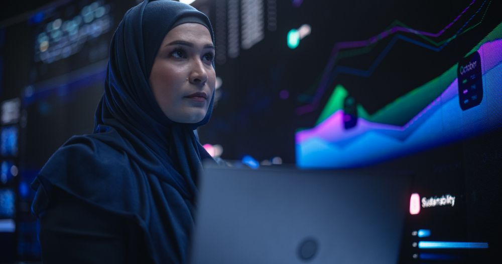 Muslim Female Data Center IT Engineer Standing in a Room with Statistics and Charts Settings on a Digital Screen.