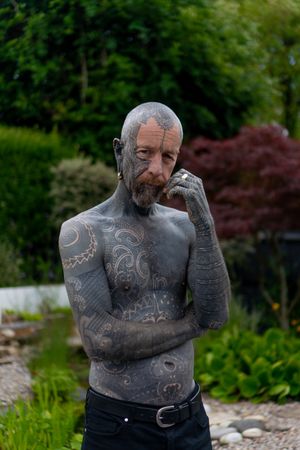 Slender bald man with extreme full-body tattoos and piercings standing in garden