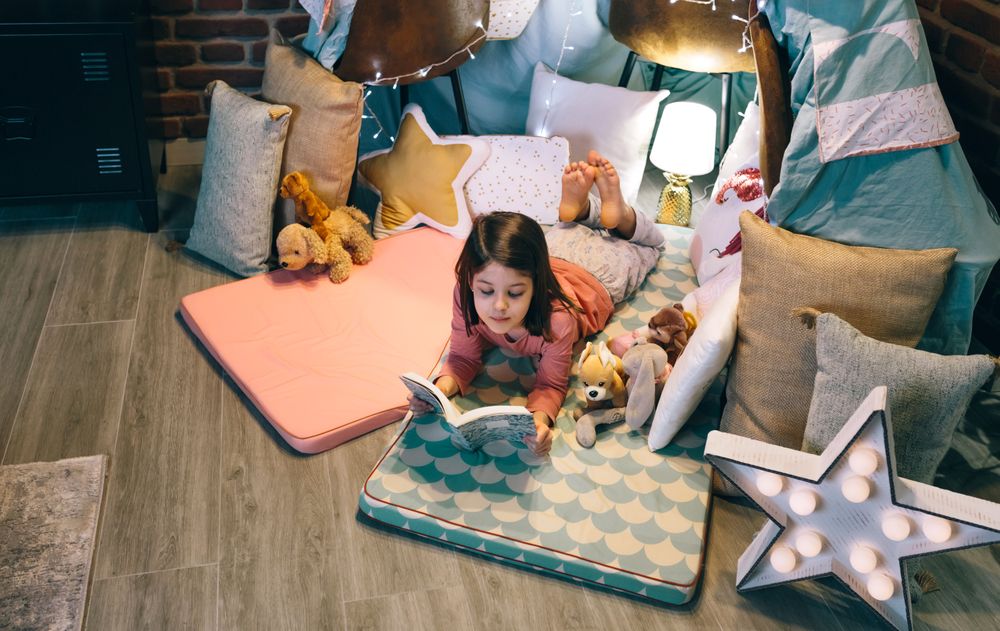 Serene little girl looking a book while resting surrounded by cushions in homemade teepee