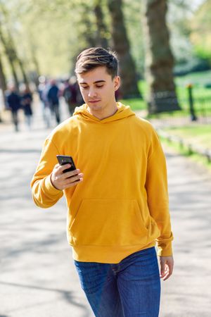 Man in yellow hoodie checking messages on cell phone while walking in park