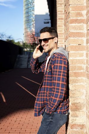 Man leaning on a brick wall wearing sunglasses and chatting on smartphone outdoors