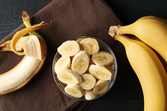 Bowl of banana slices, banana and towel on wooden background