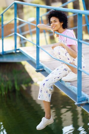 Happy woman in floral overalls sitting on bridge over water