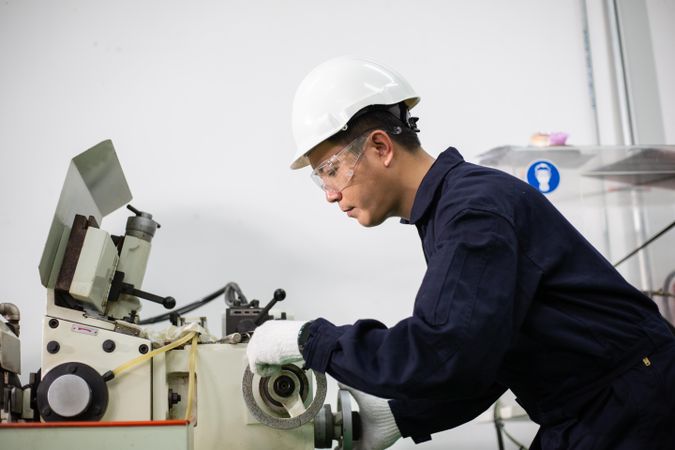 Asian male in hard hat and coveralls operating machine in factory