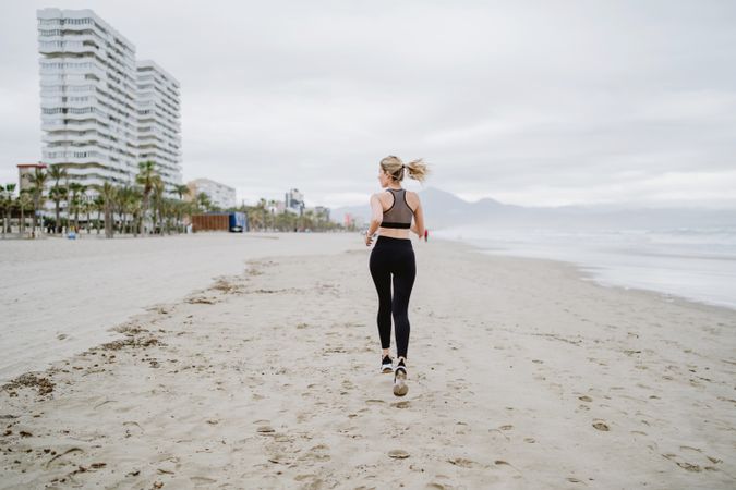 Female runner in stylish sportswear jogging along calm sandy seaside
