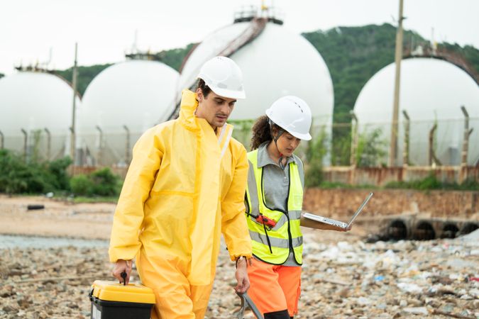 Workers in safety gear walking to survey at site near refinery storage tanks, industry facility, Hazardous environment, scientist holding toolbox, woman with laptop