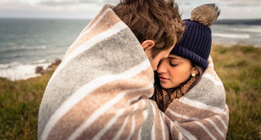 Young couple embracing under blanket in a chilly day with sea and dark cloudy sky in the background