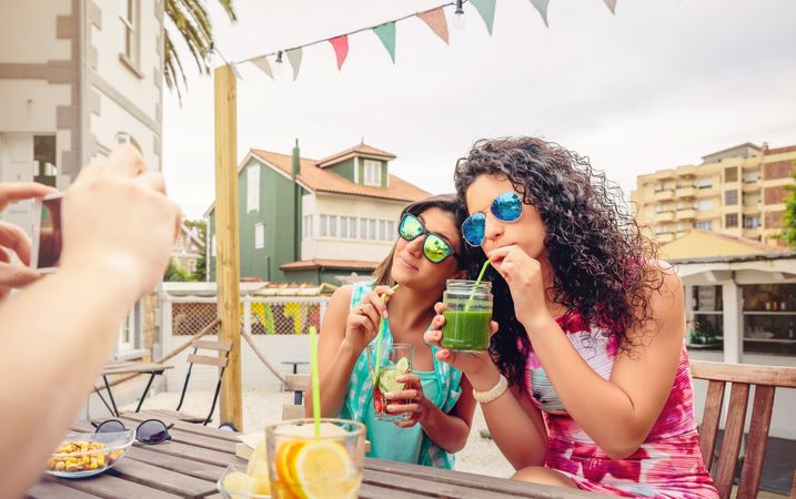 Two female friends sipping cocktails outside as friend takes photo
