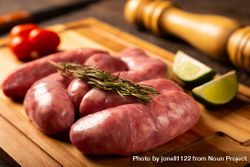 Raw sausages arranged on wooden board with rosemary, lime and tomatoes 56jVY5