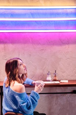 Positive woman with cup at table near wall with neon lamps