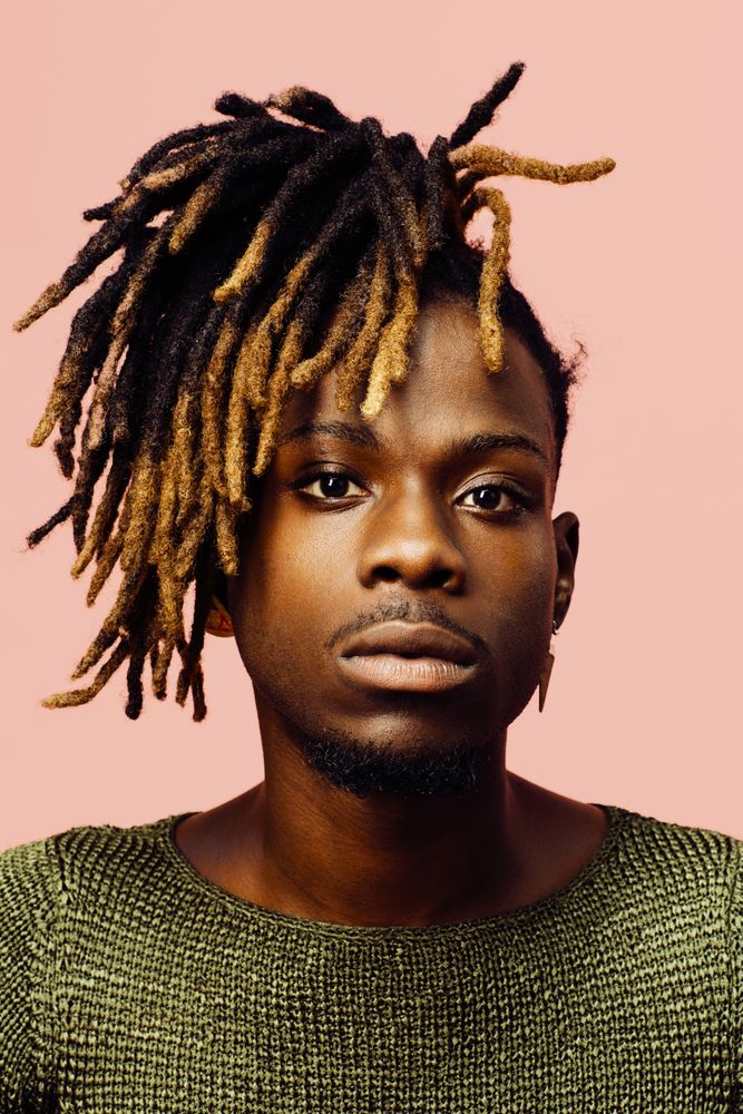 Portrait of a serious young man in green with dreadlocks and red sunglasses, isolated on pink