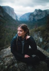 Young woman in dark bubble jacket sitting on rock formation in the woods looking away