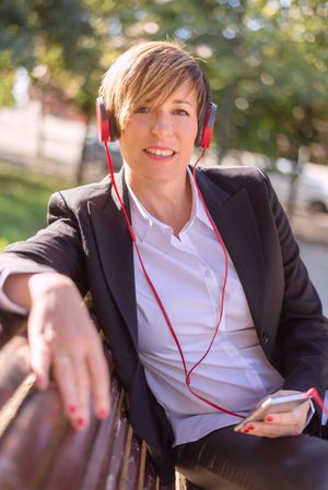 Female in blazer sitting on park bench listening to music through phone with red headphone