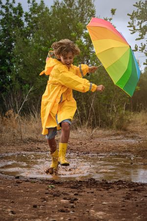 Cheerful boy with rainbow umbrella playing in puddle