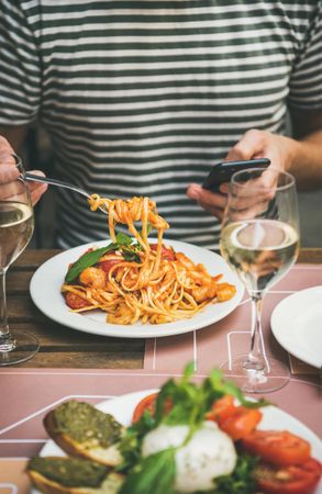 Man in striped shirt eating pasta with fork, wine, and smartphone, vertical composition